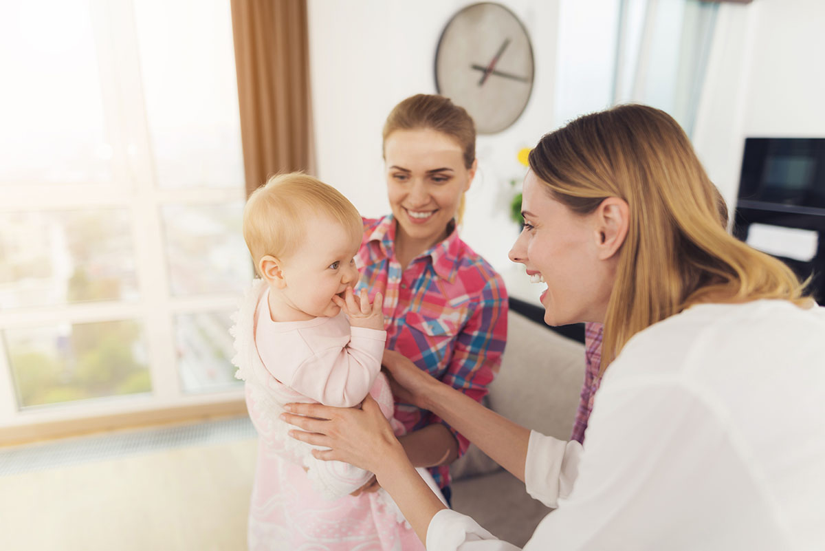 The babysitter meets the mother of the children, holding the baby in her arms. The older girl hugs Mom. They all smile. Children really like their nanny.